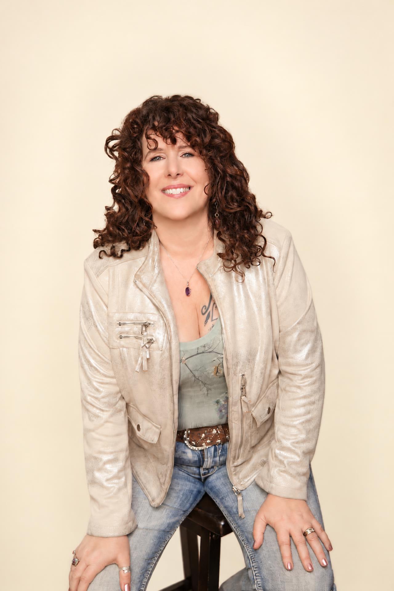 Luannah Victoria Arana seated on a stool and smiling, wearing a champagne jacket over a sage top, against a warm ivory studio backdrop